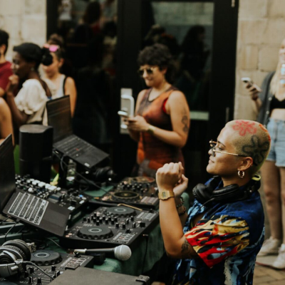 A DJ with a shaved, tattooed head and colorful shirt stands at a mixing console outdoors, performing for a crowd of people enjoying the music in front of a building with glass doors.
