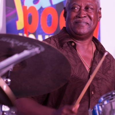 Smiling man playing drums at a jazz event, colorful jazz poster in background.