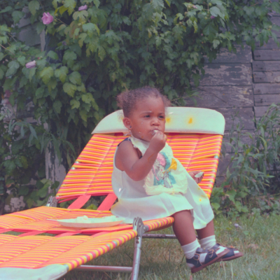 A young child wearing a light dress sits on an orange-striped lounge chair outdoors, eating something with their hand. Lush green bushes and a weathered wooden fence are in the background.