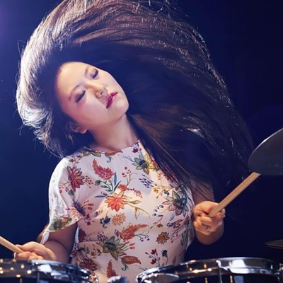 An Asian woman with long dark straight hair and wearing a white shirt with a floral pattern closes her eyes and tilts her head as she plays drums