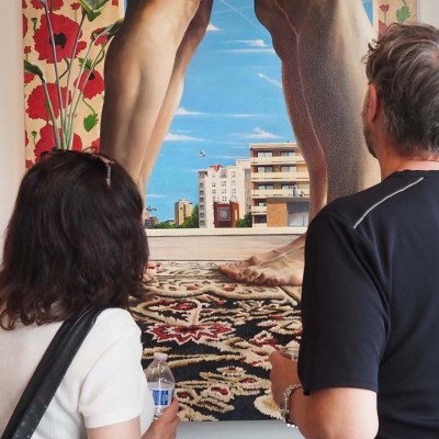 Two people stand with their back to the camera. They're looking at a large multimedia piece hanging on a gallery wall. The artwork shows two sets of bare human legs, buildings, and flowered patterns.