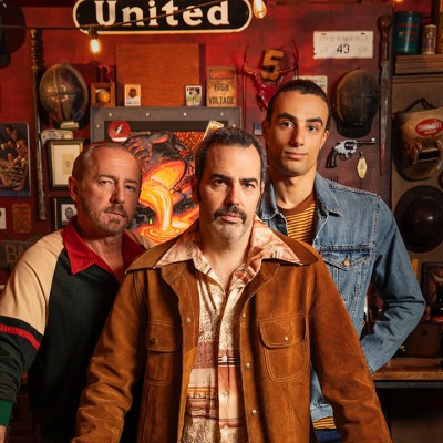 Three white male actors pose behind the counter in a junk shop, surrounded by aged items for sale
