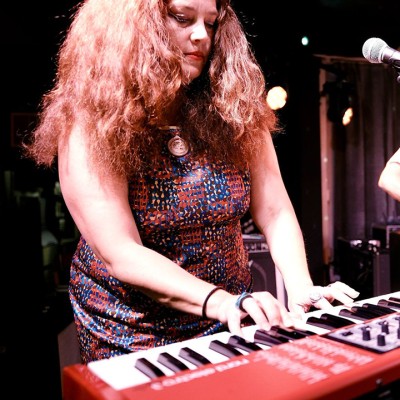 A woman with long, curly hair wearing a patterned dress plays a red keyboard on stage, focused on the keys, with stage lights in the background.