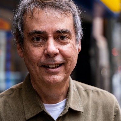 Portrait of a smiling man with short gray hair, a white T-shirt, and a light brown collared button-down shirt. He's standing outside in front of a blurred background that shows a glimpse of a theater marquee.