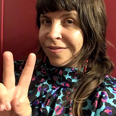 A person with long brown hair and bangs is smiling and holding up a peace sign. They are wearing a high-neck, colorful patterned top, and standing in front of a dark red background.