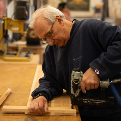 An older man uses a brad nailer to assemble wood pieces in a workshop.