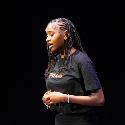 A young woman with braids speaks on stage against a black background.