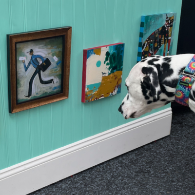 A Dalmatian sniffs a turquoise wall displaying three pieces of colorful artwork.