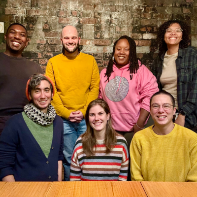 Seven people pose together in front of a brick wall, smiling at the camera.