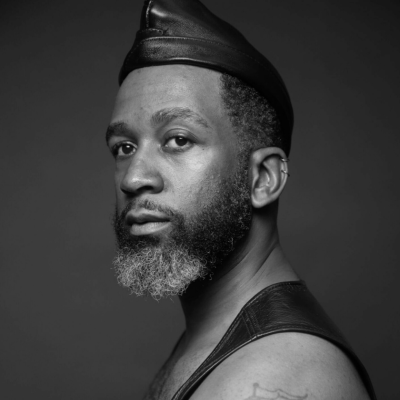 A man with a beard and leather cap looks at the camera in a black and white portrait.