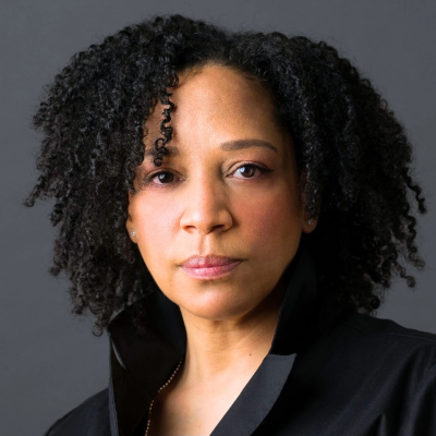 Woman with curly hair in a black shirt looks directly at the camera, neutral expression.