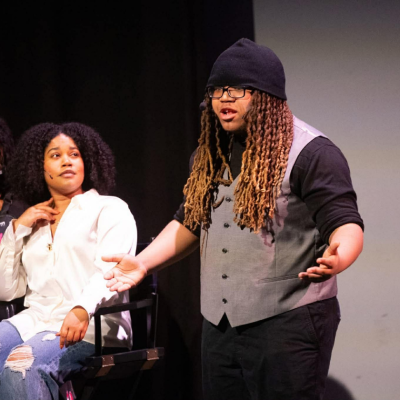 Person speaking passionately on stage as another looks on from a chair.