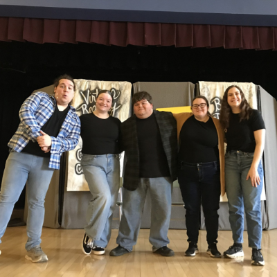 Five people stand smiling together in front of a stage backdrop, wearing casual clothes.