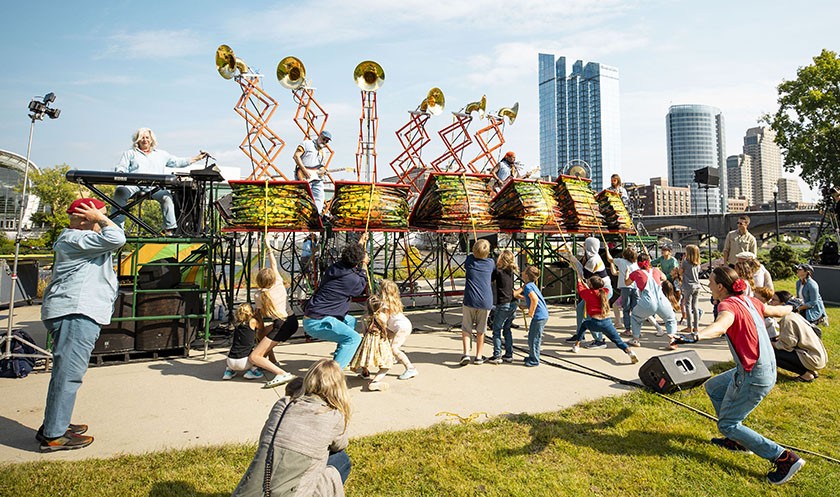 A lively outdoor performance features musicians elevated on platforms playing brass instruments, while a diverse crowd of children and adults dance and enjoy the show in a city park with tall buildings in the background.