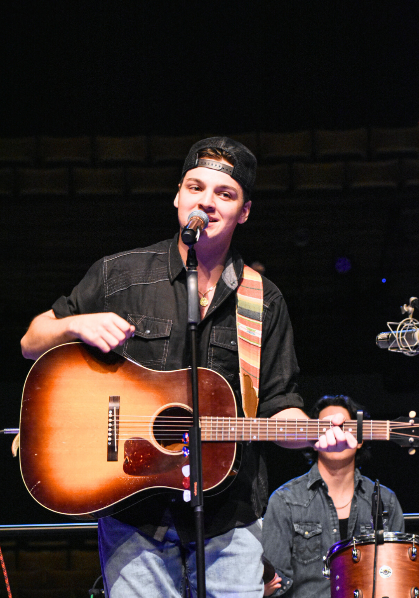 A man in a black shirt plays acoustic guitar and sings into a microphone on stage.