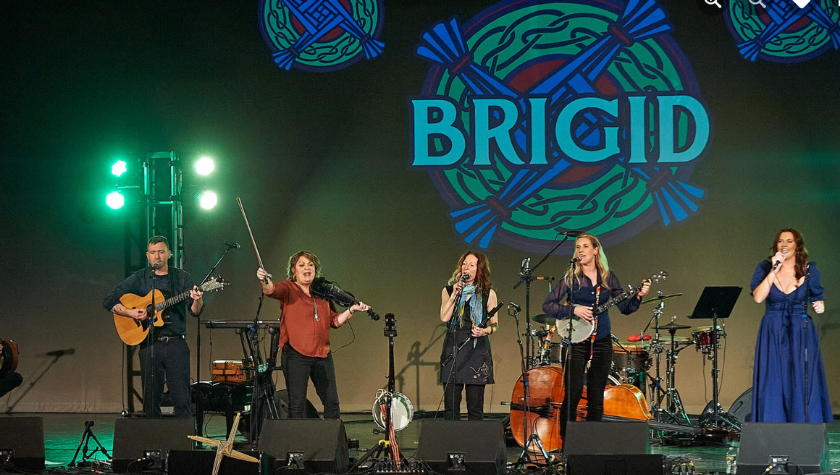Five musicians perform on stage with instruments under a BRIGID sign.