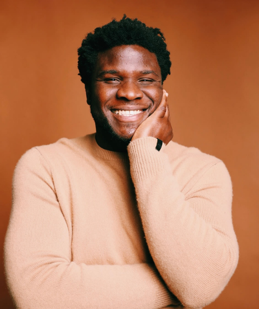 A man in a beige sweater smiles with his hand resting on his cheek.