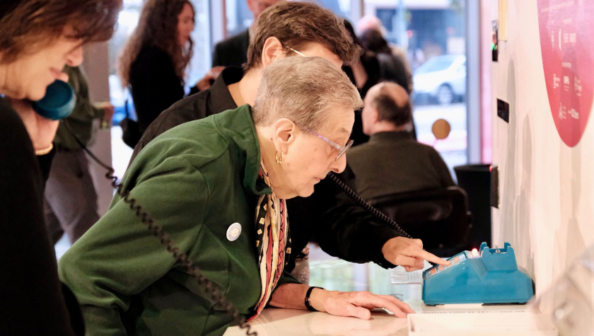 Older woman using a blue rotary phone while another person dials the number.
