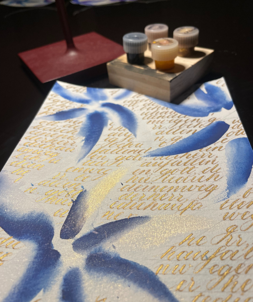 A handwritten letter with blue floral art and paint pots on a dark table.