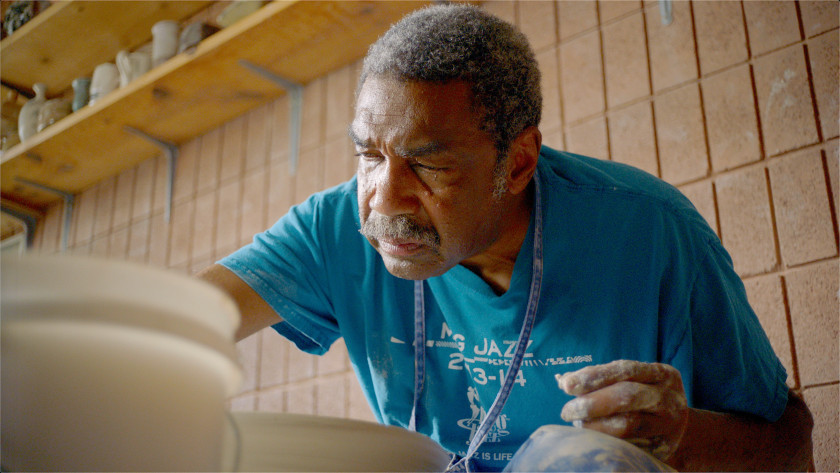 An older man with gray hair and brown skin in a blue shirt shapes pottery.