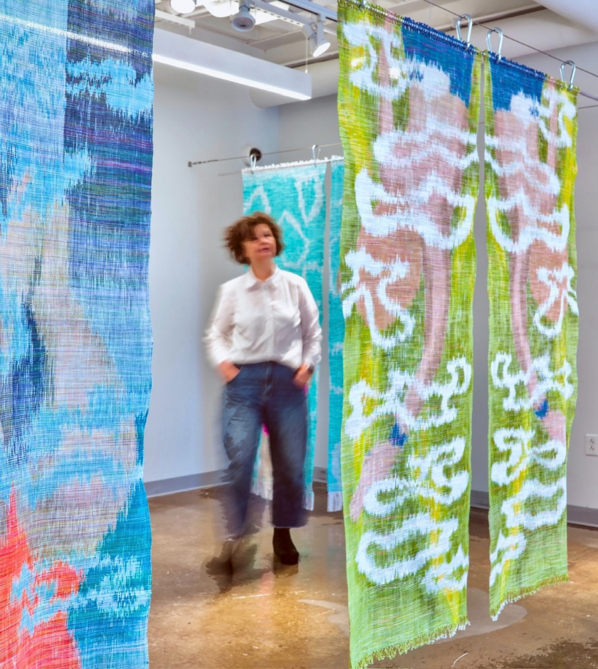 Person walking among colorful woven textile art hanging in a bright, modern gallery space.
