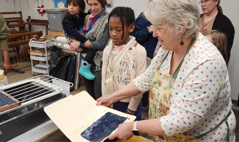 An older woman shows children how to make paper by holding a freshly made blue sheet, as a small group watches closely in a workshop setting.
