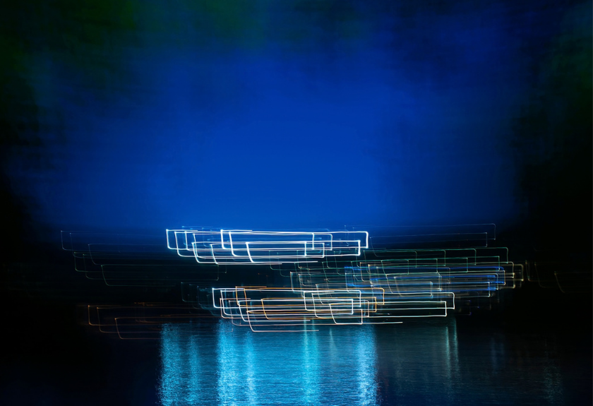 Long exposure of light trails reflected on water against a dark blue background.