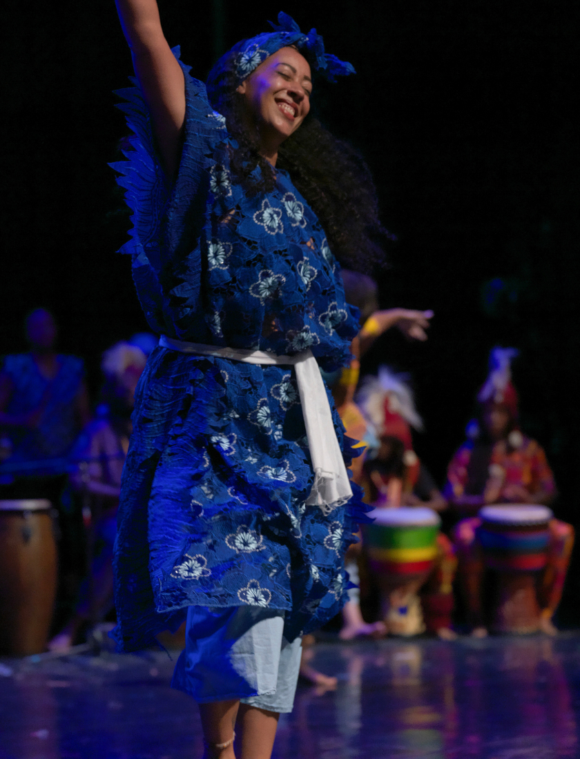 A smiling dancer in a blue lace outfit performs on stage in front of percussionists.