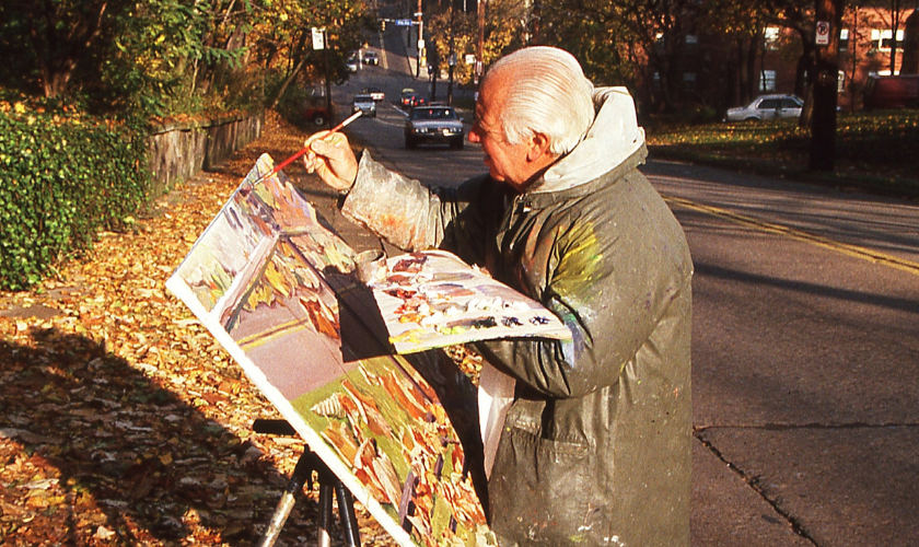 An elderly artist painting an outdoor landscape on an easel by a street in autumn.