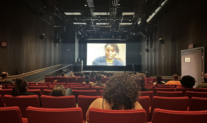 People sit in red theater seats watching a film projected on a large screen in a dark cinema. The film shows a close-up of a person speaking, with English subtitles visible at the bottom of the screen.