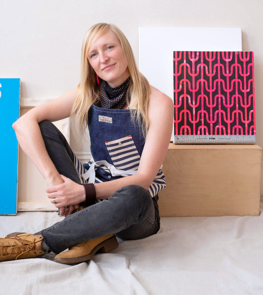 A woman in an apron sits on a floor between two pieces of art.