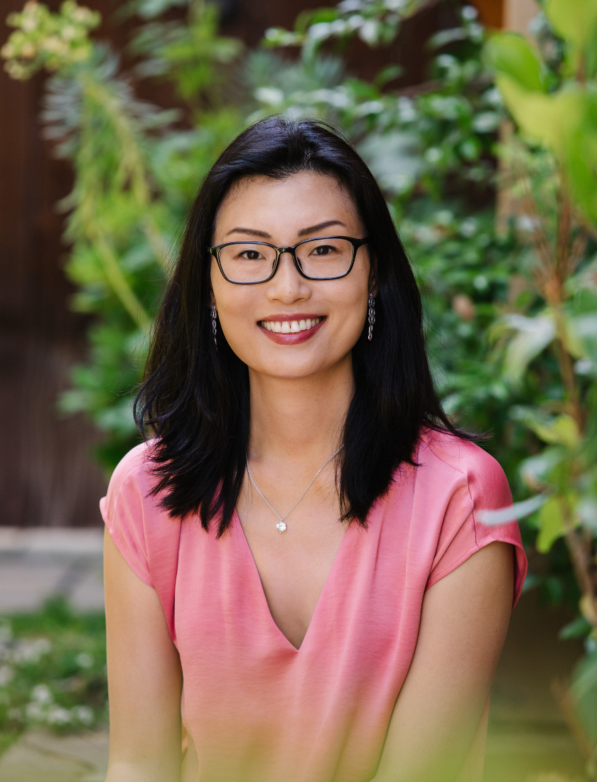 A smiling woman with dark hair and glasses wears a pink top and a silver necklace.