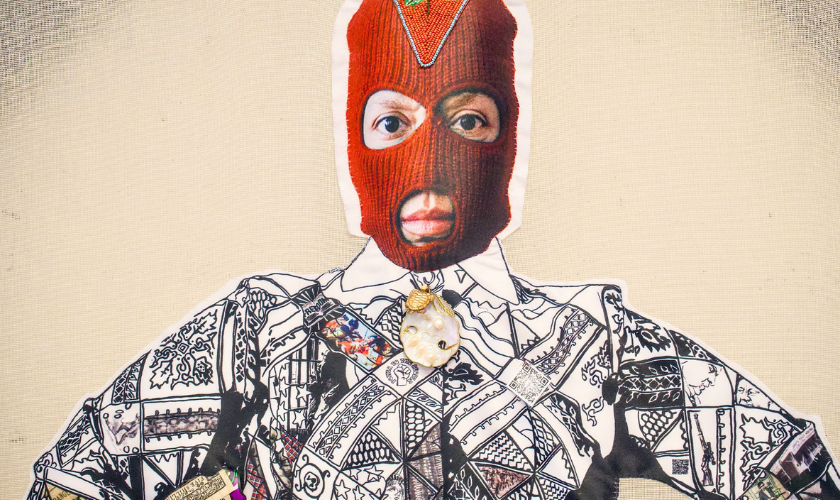 A person wearing a red balaclava exposing their eyes and mouth, dressed in a patterned black and white shirt with a large decorative brooch at the collar, standing against a light-colored background.