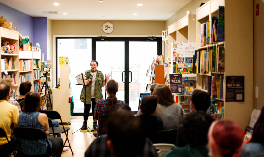 A person stands at a microphone stand reading or speaking to an audience seated in a bookstore or library, with bookshelves lining the walls and daylight coming through double glass doors in the background.