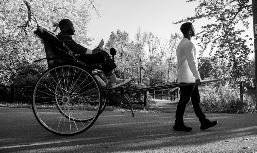A man pulls a rickshaw with a passenger holding a laptop through a park.