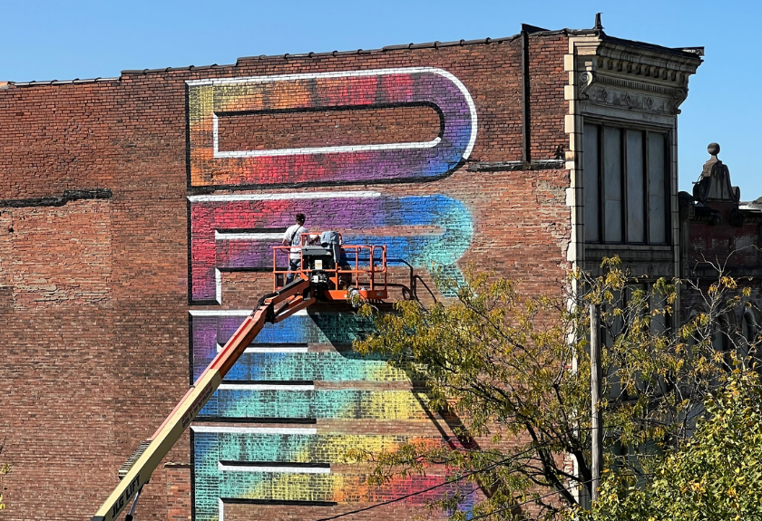 Two people in a lift paint a colorful, large-lettered mural on a brick building wall.