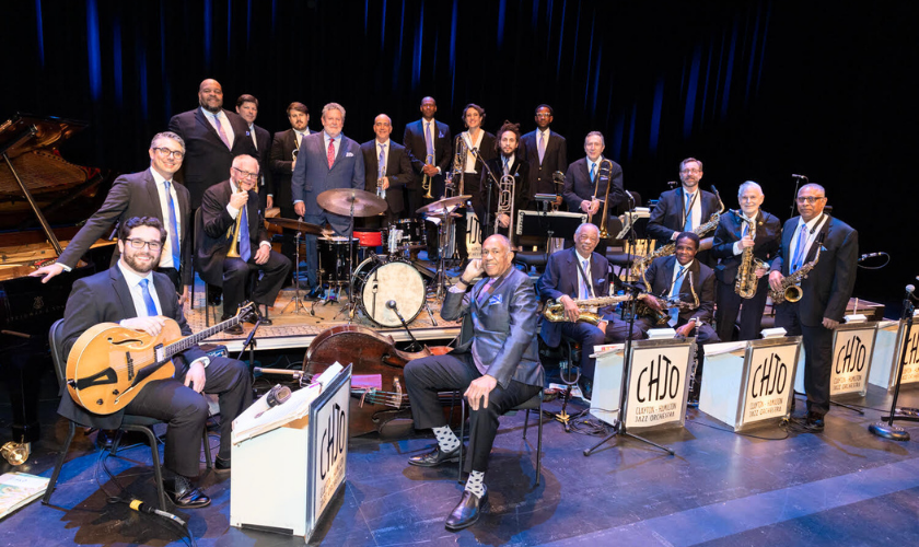 A large jazz band of musicians in suits poses with their instruments, including brass, drums, piano, guitar, and bass, on a stage with blue lighting and CHJO music stands in front.