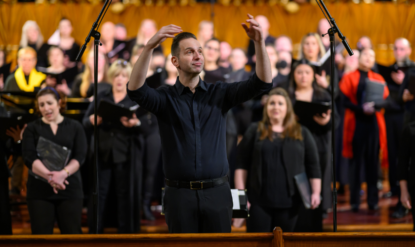 A choir conductor in black leads a group of singers who are holding sheet music. The choir stands in rows on a stage with microphones visible in front, and the setting appears to be a church or concert hall.