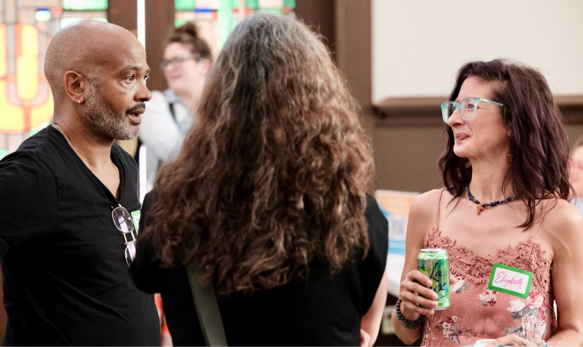 	 Three adults converse indoors at a social event. One man stands on the left, a woman with long hair is seen from behind, and another woman holding a can and wearing glasses stands on the right. Name tags are visible on two people.
