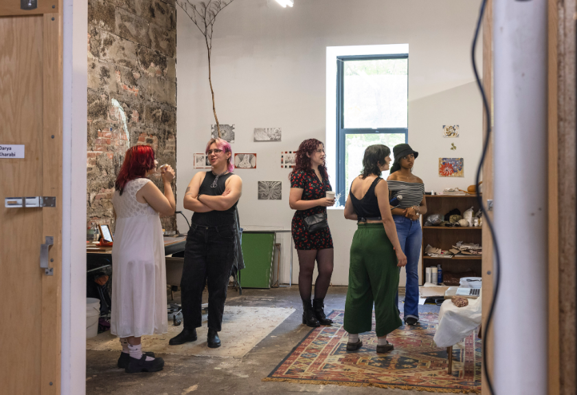 Six people stand and talk in an art studio with colorful rugs and artwork on the walls.