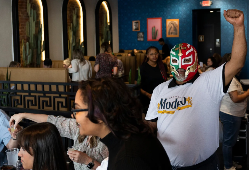 A man wearing a luchador mask and a Rodeo beer t-shirt raises his fist in a crowded restaurant.