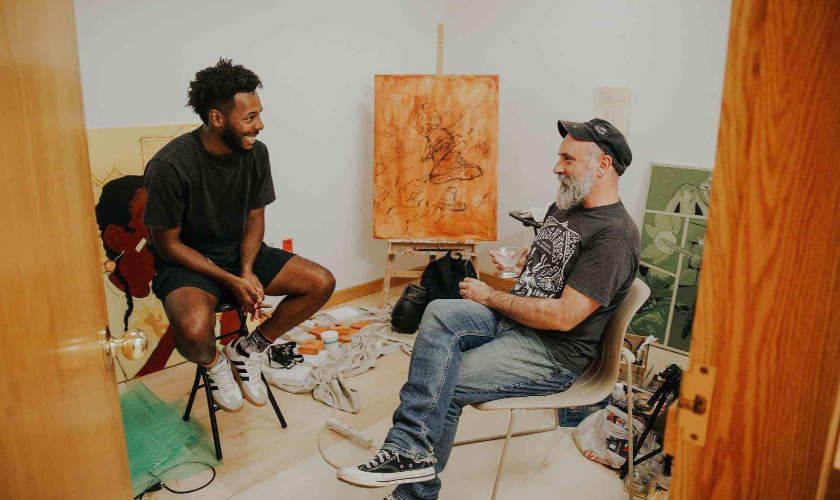 Two artists sit in a studio, smiling and talking in front of an easel with a painting.