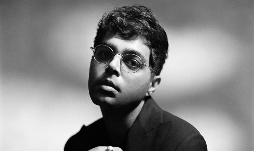 A black and white portrait of a person with short, wavy hair and round glasses, wearing a dark jacket, looking slightly upward with a neutral expression against a blurred background.