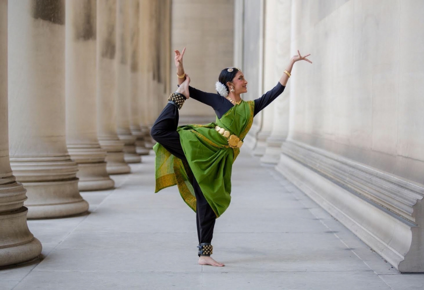 A woman in green traditional Indian attire poses in a classical dance stance between tall columns.
