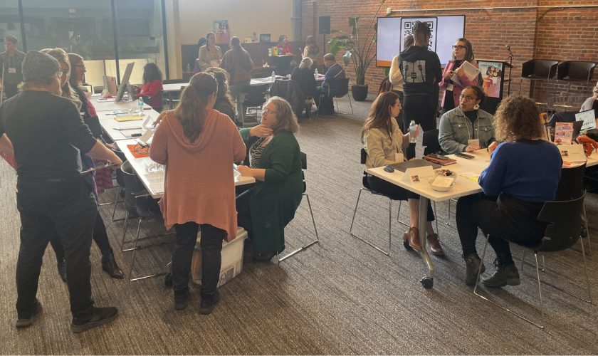 A group of people interact at various tables during an indoor event. Some sit and talk, while others stand or walk around. The space has brick walls, large windows, and informational materials on display.