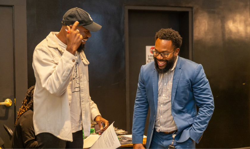 Two men smiling and laughing together indoors; one wears a cap and light jacket, and the other wears glasses and a blue blazer. They appear to be having an animated, friendly conversation.