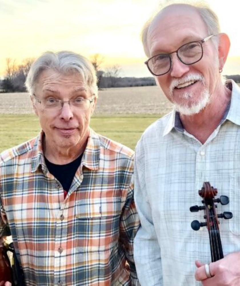 Two older men standing outside in a field at sunset, both smiling. The man on the left wears glasses and a plaid shirt. The man on the right wears glasses, a light shirt, and holds a violin.