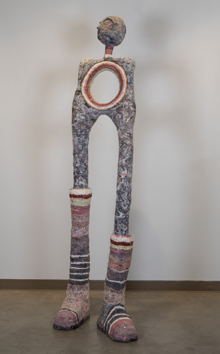 A tall, abstract humanoid sculpture with long legs, large boots, a circular hole in its torso, and a small, rounded head. The surface is textured and painted in muted colors with red, white, and purple accents.