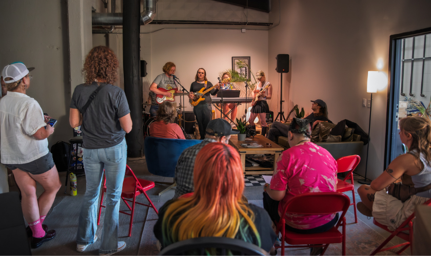 A small band performs indoors for a seated audience. People watch attentively in a cozy room with red chairs, art on the walls, and dim lighting. Some audience members stand while others sit, creating a relaxed, intimate atmosphere.