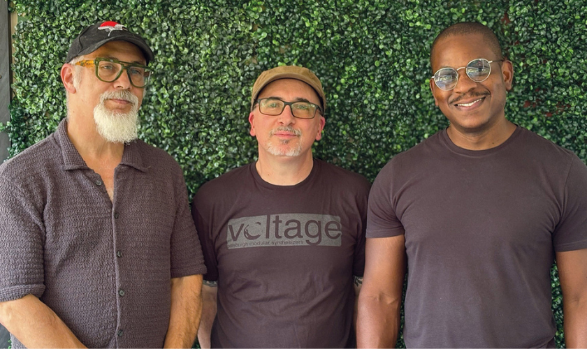 Three men stand side by side in front of a green leafy background. Two wear glasses; all are smiling. The man in the center wears a cap and a voltage t-shirt, while the others wear dark short-sleeved shirts.